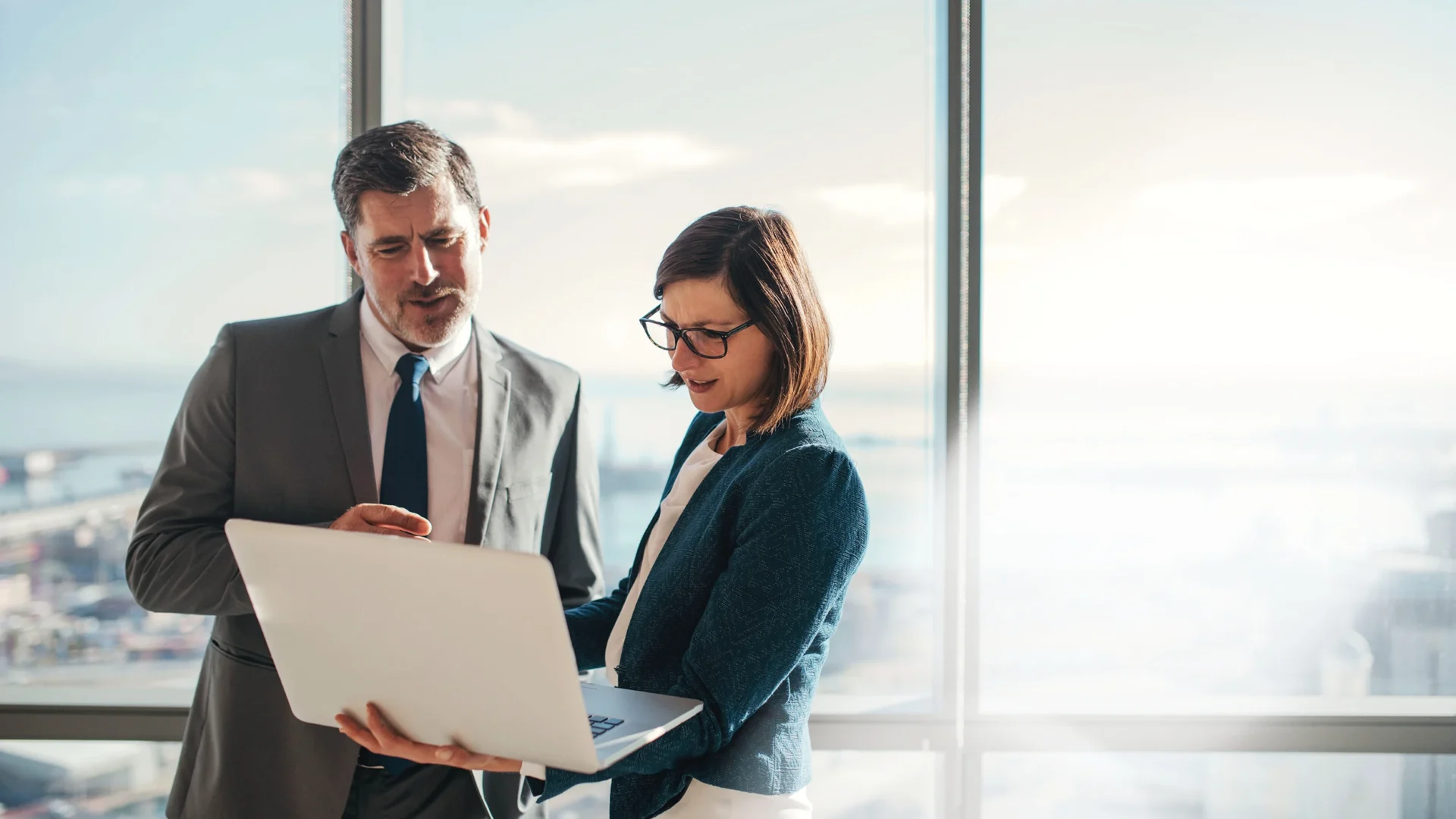 Businessman and businesswoman using a laptop together while standing in front of office building windows